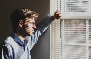 man looks out window through shades