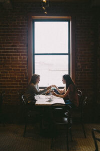 two women sit across from each other at table in front of window